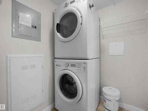 Laundry area featuring stacked front-loading appliances, a wall-mounted wire shelving unit, and tile flooring - 220 5804 Mullen Place, Edmonton, AB - Indoor Photo Showing Laundry Room