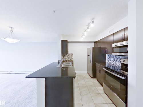 Kitchen featuring dark cabinetry, stainless steel appliances, a breakfast bar with a dark countertop, a mosaic tile backsplash, and tile flooring - 220 5804 Mullen Place, Edmonton, AB - Indoor Photo Showing Kitchen With Double Sink