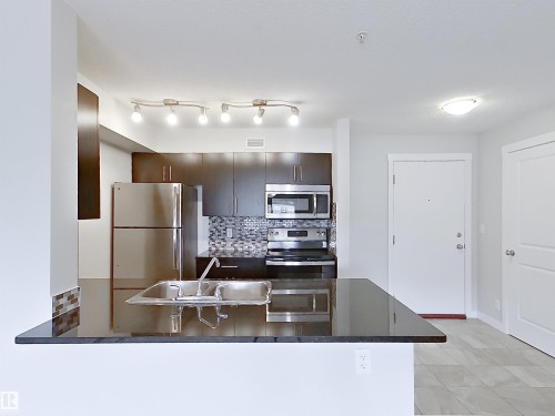 Kitchen featuring dark wood-finish cabinetry, stainless steel appliances, a mosaic tile backsplash, and a large breakfast bar with a double basin sink - 220 5804 Mullen Place, Edmonton, AB - Indoor Photo Showing Kitchen With Double Sink With Upgraded Kitchen