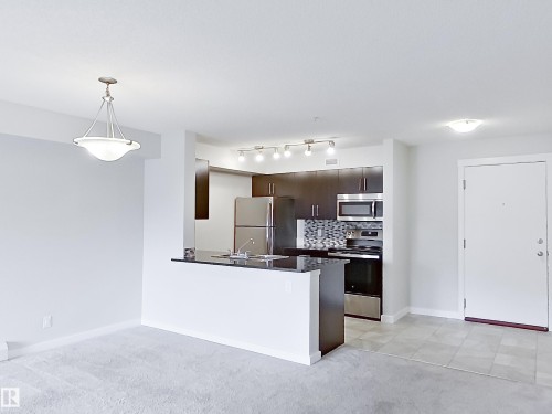 Modern kitchen featuring dark wood-finish cabinetry, stainless steel appliances, a mosaic tile backsplash, and a black countertop bar - 220 5804 Mullen Place, Edmonton, AB - Indoor Photo Showing Kitchen