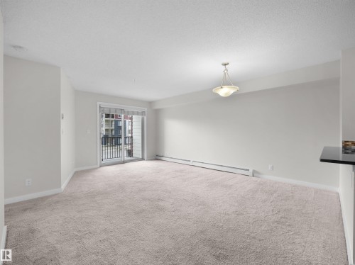 Spacious living area featuring neutral carpeting, light-toned walls, and a glass sliding door providing access to an exterior balcony - 220 5804 Mullen Place, Edmonton, AB - Indoor Photo Showing Other Room