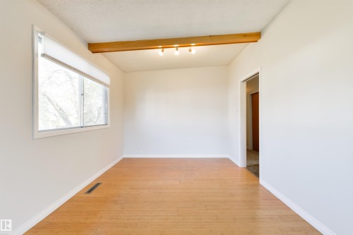 Bright interior room featuring a large window with white blinds, wood-finish flooring, a wood ceiling beam with track lighting, and a neutral wall color - 6134 37A Avenue, Edmonton, AB 
