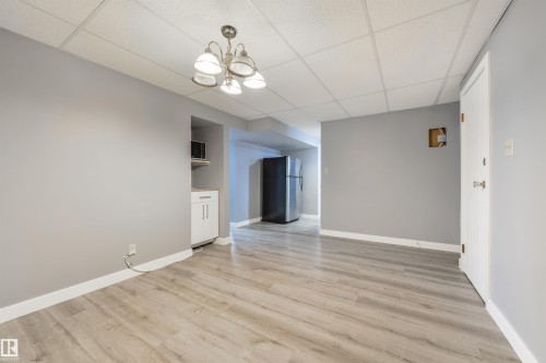 Spacious room featuring light wood-finish flooring, neutral gray walls, a drop ceiling, and a built-in cabinet with a microwave - 6134 37A Avenue, Edmonton, AB 