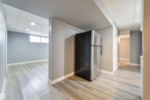 Spacious interior featuring light wood-finish flooring, neutral gray walls, a drop ceiling with recessed lighting, and a stainless steel refrigerator - 6134 37A Avenue, Edmonton, AB 