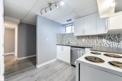 Galley kitchen featuring white cabinetry, dark countertops, mosaic tile backsplash, wood-finish flooring, and track lighting - 6134 37A Avenue, Edmonton, AB 