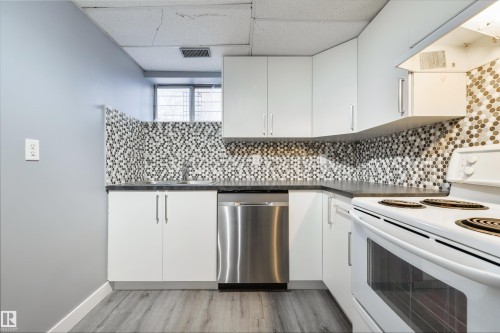 Kitchen featuring white cabinetry, dark countertops, a mosaic tile backsplash, stainless steel dishwasher, and wood-finish flooring - 6134 37A Avenue, Edmonton, AB 