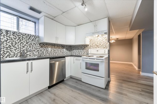 Kitchen featuring white cabinetry, dark countertops, a mosaic tile backsplash, a stainless steel dishwasher, and wood-finish flooring - 6134 37A Avenue, Edmonton, AB 