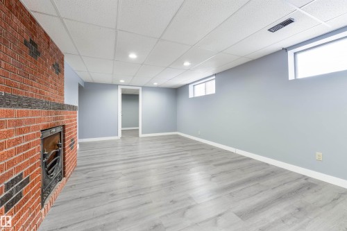 Spacious room featuring a red brick fireplace with a dark metal insert, light wood-finish flooring, recessed lighting, and two high-set windows - 6134 37A Avenue, Edmonton, AB 