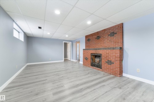 Spacious room featuring brick fireplace, light gray wood-finish flooring, drop ceiling with recessed lighting, and light blue wall tones - 6134 37A Avenue, Edmonton, AB 