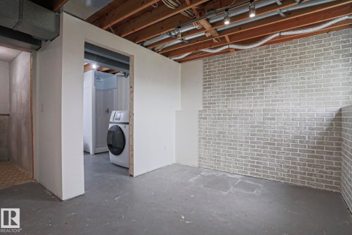 Unfinished lower level with exposed ceiling joists and ductwork, featuring painted concrete flooring, a partial brick-look accent wall, and an open doorway revealing a shower stall and laundry appliance - 35 Chungo Drive, Devon, AB - Indoor Photo Showing Other Room
