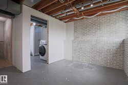 Unfinished lower level with exposed ceiling joists and ductwork, featuring painted concrete flooring, a partial brick-look accent wall, and an open doorway revealing a shower stall and laundry appliance - 