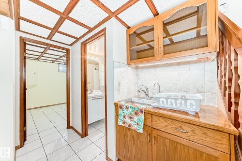 Utility area featuring a sink with a faucet, wooden cabinetry with glass-fronted upper cabinets, and a tiled backsplash - 13332 25 Street, Edmonton, AB - Indoor Photo Showing Other Room