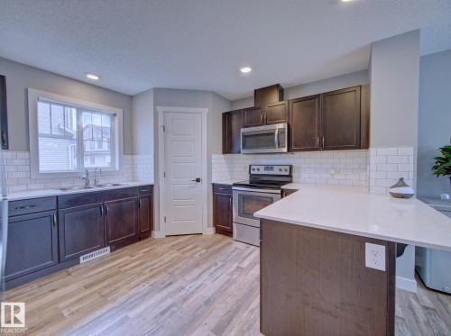 Open-concept kitchen featuring dark wood cabinetry, white subway tile backsplash, stainless steel appliances, and a central island with white countertops - 11 165 Cy Becker Boulevard, Edmonton, AB - Indoor Photo Showing Kitchen With Stainless Steel Kitchen