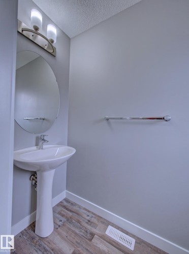 Pedestal sink with chrome faucet and circular mirror, complemented by wood-finish flooring, a chrome towel bar, and a two-light vanity fixture - 11 165 Cy Becker Boulevard, Edmonton, AB - Indoor Photo Showing Bathroom