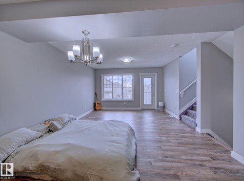 Spacious room featuring wood-finish flooring, a contemporary chandelier, large windows, a glass-paneled door, and a carpeted staircase with white railings - 11 165 Cy Becker Boulevard, Edmonton, AB - Indoor Photo Showing Bedroom