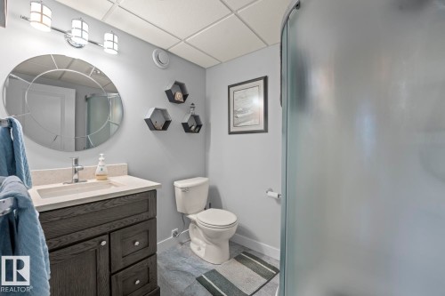 Bathroom featuring a dark wood-finish vanity with a light-toned countertop and an undermount sink, a large decorative circular mirror, a three-light vanity fixture, a toilet, and a frosted glass shower enclosure - 1304 12 Street, Cold Lake, AB - Indoor Photo Showing Bathroom