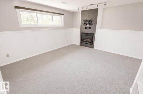 Carpeted room featuring a modern fireplace with a dark gray surround, white wainscoting with a chevron pattern, a horizontal window, and track lighting - 4510 Lakeshore Dr, St. Paul Town, AB - Indoor Photo Showing Other Room With Fireplace