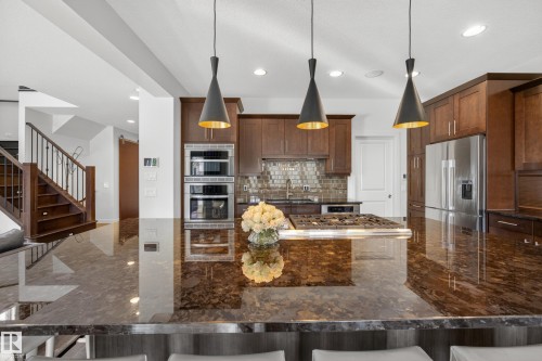 Spacious kitchen featuring an expansive island with a gas cooktop, rich wood-finish cabinetry, stainless steel appliances, and sleek pendant lighting - 2760 Watcher Way, Edmonton, AB - Indoor Photo Showing Kitchen With Upgraded Kitchen