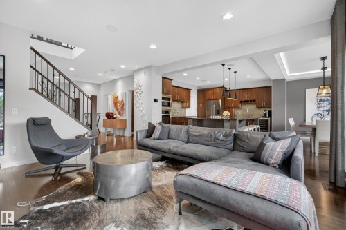 Open-concept living area featuring rich wood-finish flooring, a contemporary staircase with metal balusters, recessed lighting, a modern kitchen with wood cabinetry, and a dining area with tray ceiling detail - 2760 Watcher Way, Edmonton, AB - Indoor Photo Showing Living Room