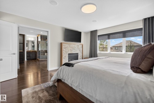 Bedroom featuring wood-finish flooring, a stone-clad fireplace, large windows with roller shades, and direct access to a bathroom with dual vanities and tiled flooring - 2760 Watcher Way, Edmonton, AB - Indoor Photo Showing Bedroom With Fireplace