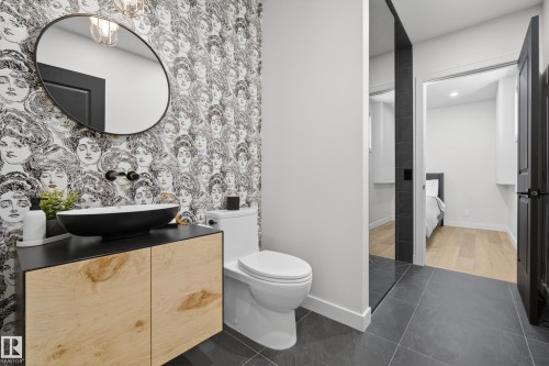 Powder room featuring a unique patterned wallpaper, a black vessel sink, a light wood-finish vanity with a black countertop, a round black-framed mirror, and a modern toilet - 2760 Watcher Way, Edmonton, AB - Indoor Photo Showing Bathroom