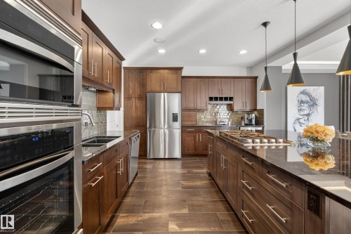 Spacious kitchen featuring rich wood cabinetry, stainless steel appliances, dark stone countertops, an integrated cooktop, and contemporary pendant lighting - 2760 Watcher Way, Edmonton, AB - Indoor Photo Showing Kitchen With Upgraded Kitchen