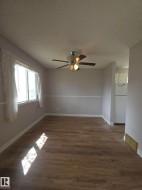 Spacious room featuring wood-finish flooring, a ceiling fan with integrated lighting, and a large window with white trim - 