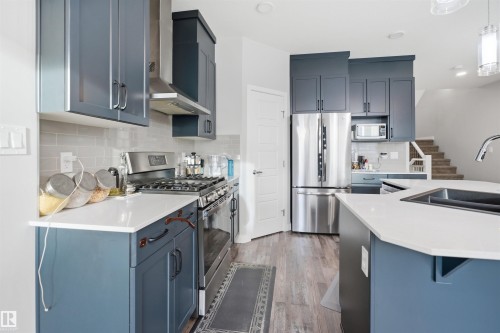Contemporary kitchen featuring blue-gray cabinetry, white countertops, stainless steel appliances, a subway tile backsplash, and wood-finish flooring - 2284 Chokecherry Close, Edmonton, AB - Indoor Photo Showing Kitchen With Double Sink With Upgraded Kitchen