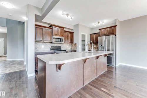 Kitchen featuring dark wood cabinetry, stainless steel appliances, a tile backsplash, and an island with a built-in sink and light-toned countertop - 2138 32B Street, Edmonton, AB - Indoor Photo Showing Kitchen With Stainless Steel Kitchen With Upgraded Kitchen