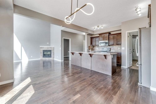 Open-concept living area featuring rich wood-finish flooring, a contemporary fireplace with a tiled surround, a large kitchen island with a breakfast bar, dark wood cabinetry, and stainless steel appliances - 2138 32B Street, Edmonton, AB - Indoor Photo Showing Kitchen
