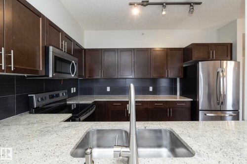 Contemporary kitchen featuring granite countertops, dark wood cabinetry with brushed nickel hardware, a stainless steel undermount sink with a high-arc faucet, stainless steel appliances, and a dark tile backsplash - 3104 10152 104 Street, Edmonton, AB - Indoor Photo Showing Kitchen With Stainless Steel Kitchen With Double Sink With Upgraded Kitchen