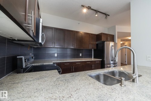 Kitchen featuring light-toned countertops, a double basin stainless steel sink with a high-arc faucet, dark wood-finish cabinetry, and a dark tile backsplash - 3104 10152 104 Street, Edmonton, AB - Indoor Photo Showing Kitchen With Stainless Steel Kitchen With Double Sink