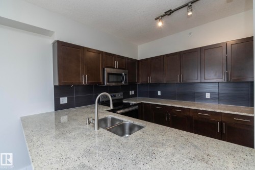 Kitchen featuring light-toned countertops, dark wood cabinetry, a stainless steel double basin sink with a high-arc faucet, and a black tile backsplash - 3104 10152 104 Street, Edmonton, AB - Indoor Photo Showing Kitchen With Double Sink With Upgraded Kitchen