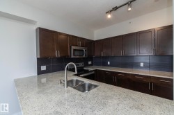 Kitchen featuring light-toned countertops, dark wood cabinetry, a stainless steel double basin sink with a high-arc faucet, and a black tile backsplash - 