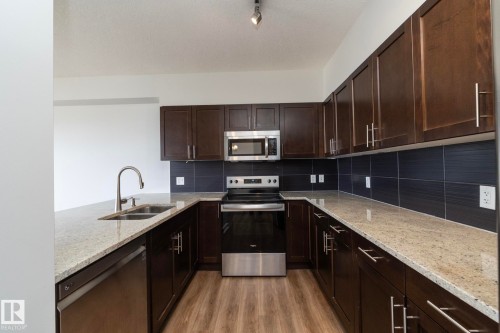 Kitchen featuring dark wood-finish cabinetry, light speckled countertops, a stainless steel sink with a gooseneck faucet, a stainless steel dishwasher, and a stainless steel range with a microwave above - 3104 10152 104 Street, Edmonton, AB - Indoor Photo Showing Kitchen With Stainless Steel Kitchen With Double Sink
