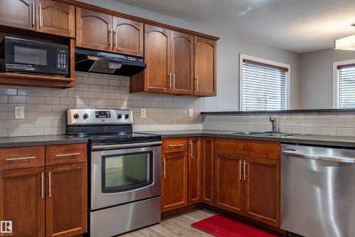 Kitchen featuring rich wood cabinetry, stainless steel appliances, dark countertops, a subway tile backsplash, and wood-finish flooring - 17715 6 Avenue, Edmonton, AB - Indoor Photo Showing Kitchen With Stainless Steel Kitchen