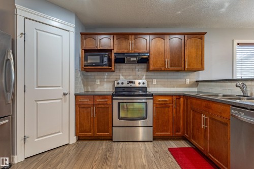 Kitchen featuring wood-finish flooring, rich wood cabinetry, stainless steel appliances, dark countertops, and a subway tile backsplash - 17715 6 Avenue, Edmonton, AB - Indoor Photo Showing Kitchen With Stainless Steel Kitchen With Double Sink