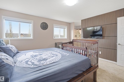 Bedroom featuring light beige walls, two windows with white trim, a textured accent wall with vertical wood-finish panels, and soft carpeting - 1444 37C Avenue, Edmonton, AB - Indoor Photo Showing Bedroom
