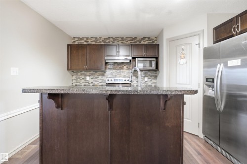Kitchen featuring an island with a speckled countertop, dark wood-finish cabinetry, stainless steel appliances, a mosaic tile backsplash, and wood-finish flooring - 1144 35 Avenue, Edmonton, AB - Indoor Photo Showing Kitchen With Upgraded Kitchen