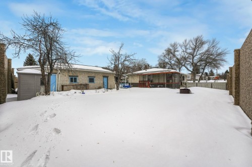 Expansive snow-covered backyard featuring a detached accessory structure with stone-look siding and vibrant blue doors, a screened-in deck with dark wood-finish railing, a picket fence perimeter, and mature trees - 2 Scarboro Place, St. Albert, AB - Outdoor