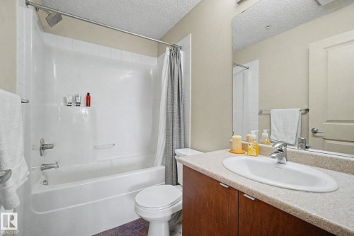 Bathroom featuring a combination bathtub and shower with a white surround, a toilet, a vanity with a light-colored countertop, an undermount sink, and a large mirror - 334 504 Albany Way, Edmonton, AB - Indoor Photo Showing Bathroom