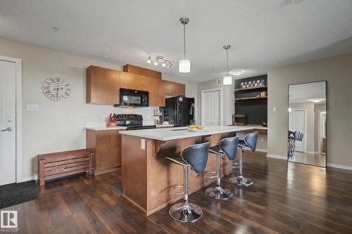Kitchen featuring wood-finish flooring, a central island with countertop seating, white countertops, built-in cabinetry, and a full suite of appliances - 334 504 Albany Way, Edmonton, AB - Indoor Photo Showing Kitchen