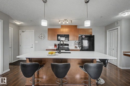 Kitchen featuring a large central island with white countertop, pendant lighting, wood-finish flooring, and light wood cabinetry with black appliances - 334 504 Albany Way, Edmonton, AB - Indoor Photo Showing Kitchen