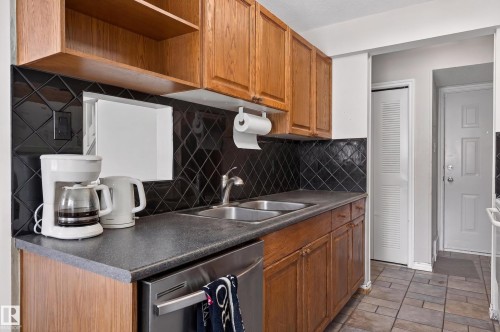 Kitchen featuring wood cabinetry, dark countertops, and a black diamond-patterned tile backsplash - 26E Meadowlark Village, Edmonton, AB - Indoor Photo Showing Kitchen With Double Sink