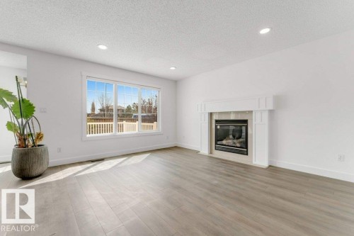 Living area with a built-in fireplace featuring a white mantel, wood-finish flooring, a large multi-pane window, and recessed ceiling lighting - 7403 Singer Landing Landing, Edmonton, AB - Indoor Photo Showing Living Room With Fireplace