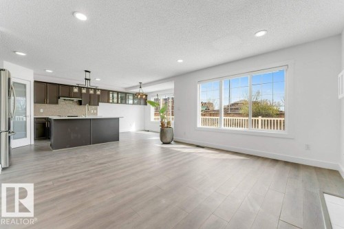 Open-concept living space featuring wood-finish flooring, recessed lighting, expansive windows, a kitchen island with sink, and dark cabinetry - 7403 Singer Landing Landing, Edmonton, AB - Indoor