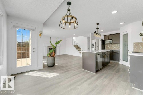 Open-concept living area with light wood-finish flooring, a contemporary kitchen island featuring an undermount sink, dark cabinetry, and stone-look backsplash, and a white fireplace mantel - 7403 Singer Landing Landing, Edmonton, AB - Indoor Photo Showing Kitchen With Upgraded Kitchen
