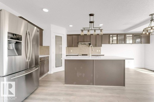 Contemporary kitchen featuring a large central island with a white countertop, stainless steel refrigerator, dark wood-finish cabinetry, geometric tile backsplash, and recessed lighting - 7403 Singer Landing Landing, Edmonton, AB - Indoor Photo Showing Kitchen With Double Sink