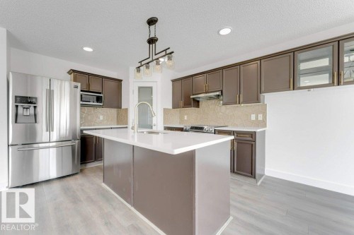Contemporary kitchen featuring a center island with a sink, stainless steel appliances, dark wood cabinetry, light wood-finish flooring, and a decorative tile backsplash - 7403 Singer Landing Landing, Edmonton, AB - Indoor Photo Showing Kitchen With Upgraded Kitchen