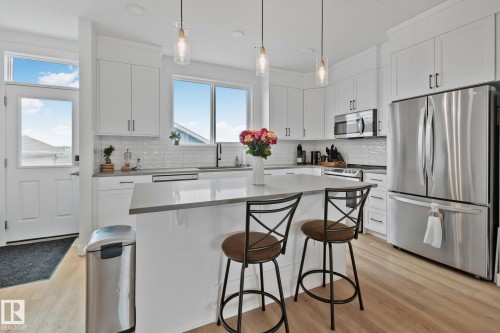 Modern kitchen featuring an expansive center island with seating, white shaker cabinetry, stainless steel appliances, and wood-finish flooring - 21027 130 Avenue, Edmonton, AB - Indoor Photo Showing Kitchen With Upgraded Kitchen